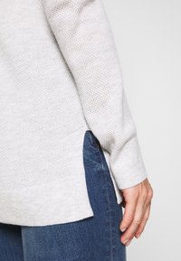 Pull en tricot gris clair à manches longues, tissu texturé, fentes sur les côtés à l'ourlet, porté sur un jean bleu foncé avec un léger délavage.