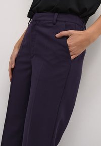 Pantalons violets avec une texture lisse, dotés d'une poche latérale et d'une coupe ajustée. La taille est finie avec des passants de ceinture.