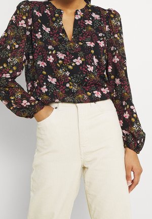Femme portant un chemisier noir à manches longues à fleurs roses et jaunes, rentré dans un pantalon en velours côtelé crème taille haute, main dans la poche.