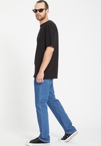 Camiseta de algodón negra con un pequeño logo rojo, combinada con jeans de pierna recta azules y zapatillas negras de corte bajo. Gafas de sol claras completan el look.