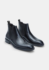 Botas Chelsea de cuero negro con paneles laterales elásticos y tiradores. Presentan un diseño elegante, puntera afilada y una textura suave y brillante.