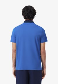 Blå polo skjorta med korta ärmar, med en kontrasterande krage och 'LACOSTE'-logotyp på baksidan. Mjuk tyg med en slät textur.