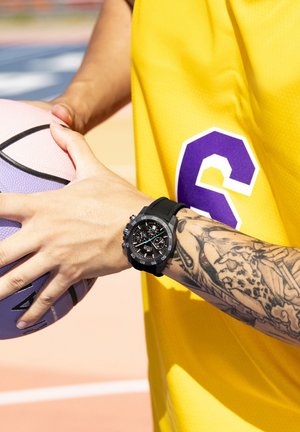 Montre de sport noire avec un bracelet en caoutchouc texturé, dotée d'un cadran rond avec plusieurs compteurs, portée sur un poignet tatoué, tenant un ballon de basket violet.