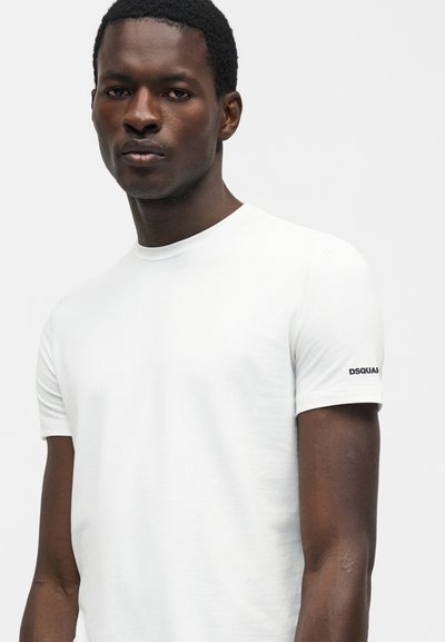 Νέος μαύρος άντρας φοράει ένα απλό λευκό T-shirt με το λογότυπο κειμένου "DSQUARED2" στο αριστερό μανίκι, πάνω σε απλό ανοιχτό φόντο.