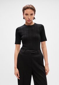 Juoda raukinukų trumpomis rankovėmis palaidinė su apvaliu iškirpimu, turinti prigludimą ir šiek tiek blizgesio, derinama su juodomis pritaikytomis kelnėmis.