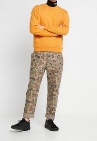 Oranje sweatshirt met geribde manchetten en zoom. Khaki camo-broeken met een relaxte pasvorm en opgerolde pijpen. Zwarte sneakers met gestructureerde zolen.