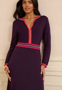 Abito lungo viola a maniche lunghe con scollatura a V, caratterizzato da bordo ondulato rosa attorno al collo e alla vita, e dettagli dorati sui polsini.