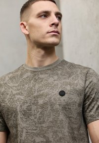 Krátké tričko s krátkým rukávem olivově zelené barvy, vyrobené z texturované látky s jemným abstraktním vzorem a černou nášivkou s logem na hrudi.