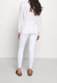 Blusa blanca de manga larga, de tejido suave, con detalles en la costura de la parte trasera, combinada con jeans skinny blancos y sandalias planas marrones. Fondo liso.