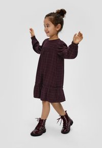 Bordeauxfarbenes Kleid mit Puffärmeln, einem ausgeflogenen Saum und einem goldenen Rautenmuster, kombiniert mit glänzenden burgunderfarbenen Schnürstiefeln.