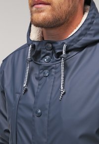 Navyblå vandtæt jakke med blødt fleecefor, justerbar hætte med snøre og kontrastfarvede snore. Har plastikknapper og en brystlomme.