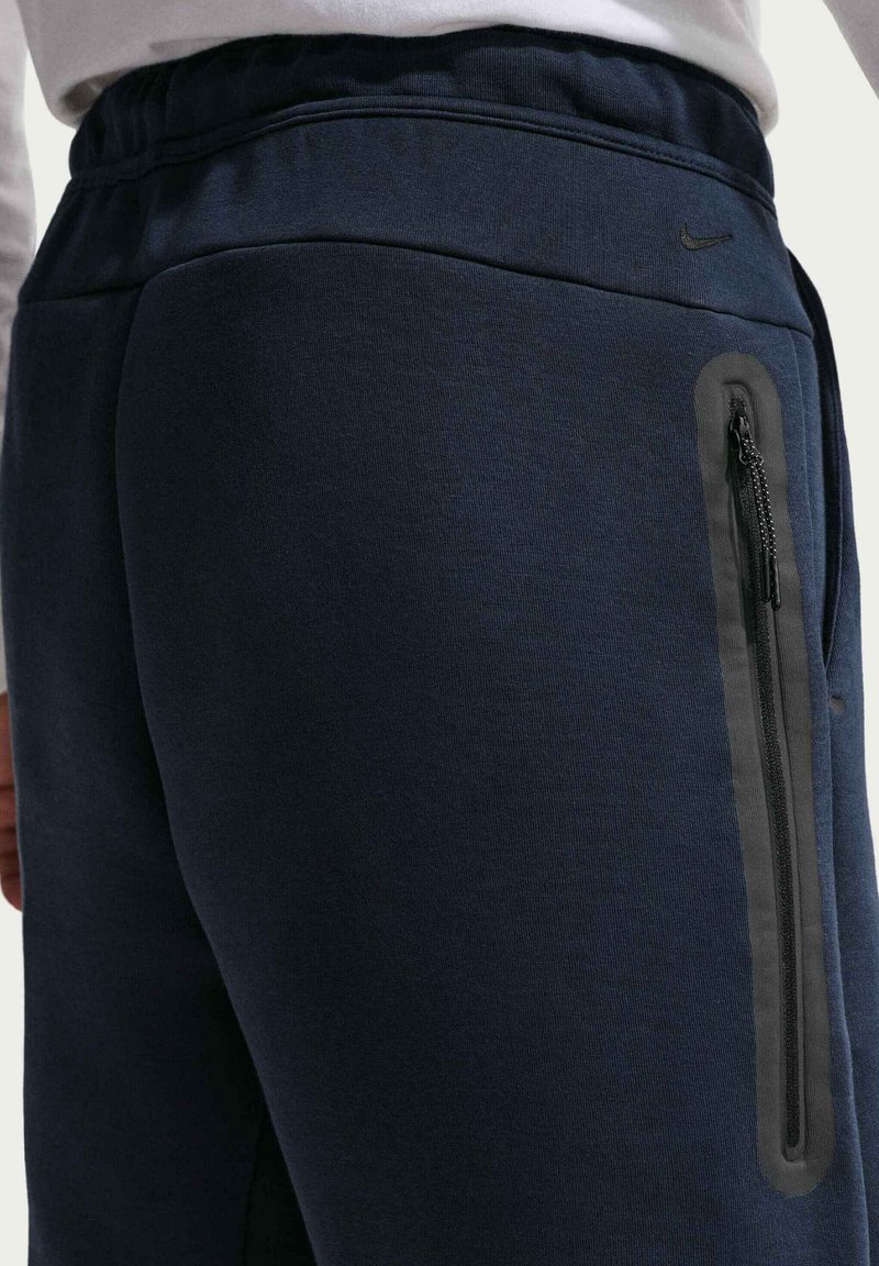 Persoon draagt donkerblauwe Nike joggingbroek met elastische tailleband en een verticale ritszak aan de zijkant, gezien vanaf de taille naar beneden.