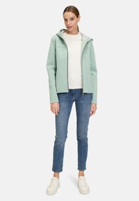 Miętowa bluza z kapturem na zamek, wyłożona szarym materiałem, połączona z kremową bluzką i niebieskimi dżinsami. Model ma na sobie białe sneakersy.