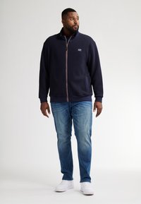 Marinblå fleecejacka med ett texturerat mönster, hel dragkedja och hög krage, kombinerad med ljusblå jeans och vita sneakers.