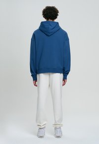 Felpa blu in cotone con vestibilità rilassata, cappuccio ampio e polsini a coste. Abbinata a pantaloni della tuta bianchi e sneakers chiare.