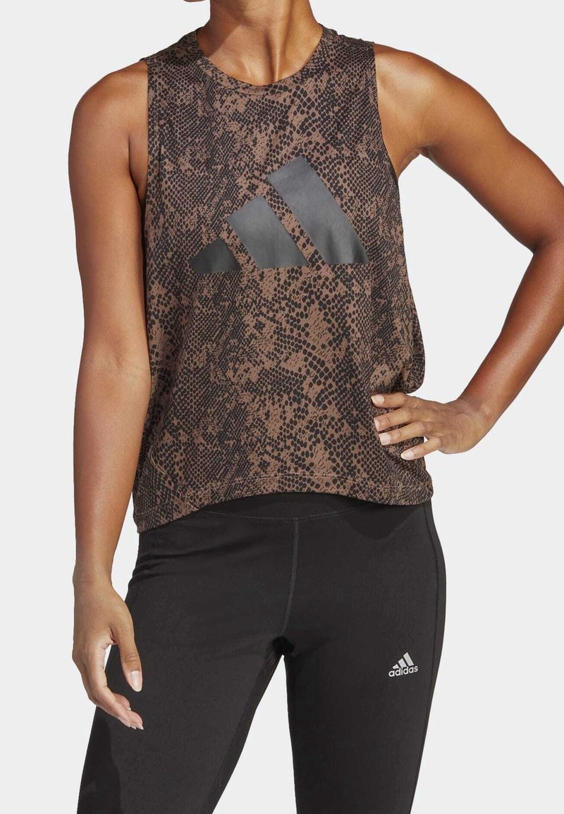 Top atlético sin mangas de color marrón con un patrón texturizado y logotipo de Adidas en negro. Combinado con leggings ajustados negros que presentan un acento de logotipo.