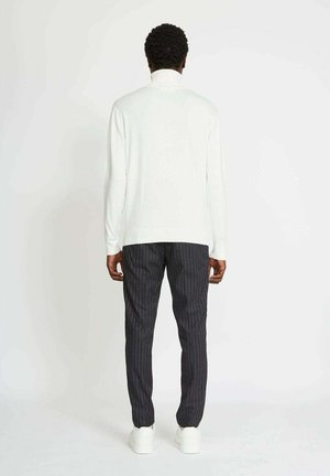 Top bianco a maniche lunghe, vestibilità comoda, abbinato a pantaloni scuri a righe. Il modello è in piedi con la schiena rivolta verso la fotocamera, indossando sneaker bianche.