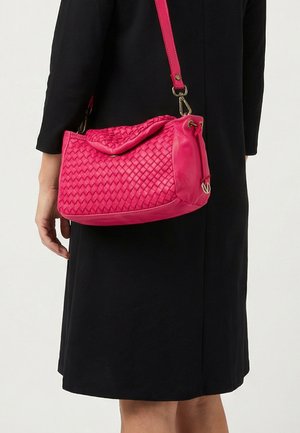 Vrouw gekleed in een zwarte jurk draagt een roze gevlochten lederen schoudertas met klep en verstelbare riem.