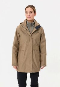 Beige regenjas met een capuchon, twee voorzakken en een gevoerde binnenlaag met rits. Heeft een donkerblauwe accent op de schouder. Gladde textuur.