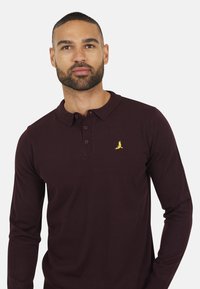 Polo a maniche lunghe color bordeaux con una finitura texturizzata, dotato di una placca a tre bottoni e un piccolo logo di un uccello ricamato giallo.