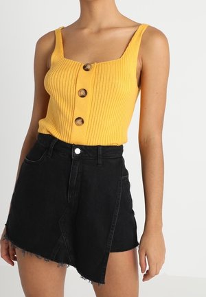 Vrouw draagt een geel geribd mouwloos topje met drie bruine knopen en een zwarte asymmetrische spijkerrok met rafelige zoom.