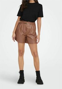 Brązowe szorty z ekoskóry z wysokim stanem, akcentami paska i plisami, w połączeniu z czarnym t-shirtem z krótkim rękawem oraz czarnymi butami wojskowymi.