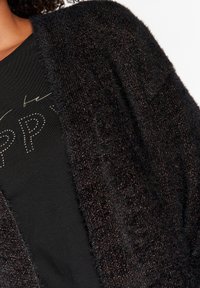 Svart, fluffig cardigan med metallisk bronsdetaljer, med öppen framsidan och texturerad yta. Lagd över en mörk t-shirt med tryckt text.