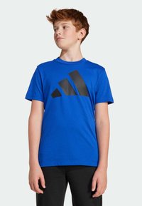 Mėlynas medvilnės marškinėlis su dideliu juodu Adidas logotipu geometriniu dizainu. Apvalus kaklas, trumpomis rankovėmis ir laisvu siluetu.