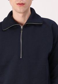 Sudadera de cremallera azul marino con un alto cuello, con cremallera plateada y un sutil logo de llamas en el lado izquierdo del pecho. Fabricada con una suave tela texturizada.