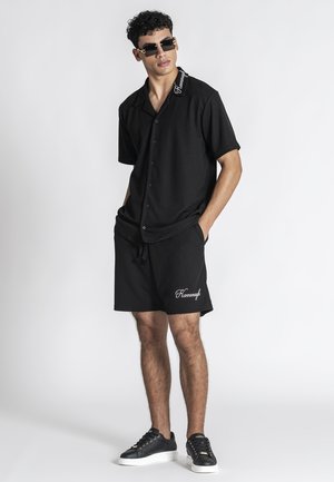 Ung mand iført sort kortærmet skjorte med knapper og shorts med hvidt tryk, sorte solbriller og sorte sneakers med hvide såler.