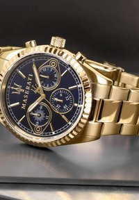 Guld-tonet Maserati ur med en sort skive, tre undermålinger og en tekstureret bezel. Har et metalarmbånd og fremtrædende timeindekser.