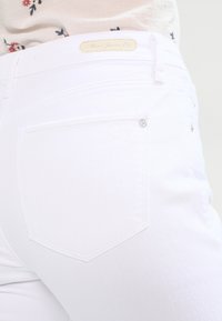 Jeans de mezclilla blanca que presentan una vista trasera, mostrando un parche rectangular encima de la cinturilla y un bolsillo trasero con un detalle de botón metálico.