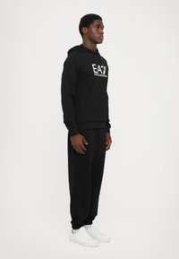 Felpa nera con tasca frontale e logo bianco. Abbinata a pantaloni jogger neri e sneakers bianche. Design semplice e aderente, con texture liscia.