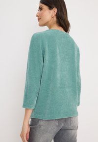 Tygsweater i teal med ribbat mönster, 3/4-långa ärmar och rundad halsringning, matchad med grå, något slitna jeans. Slät textur, avslappnad passform.
