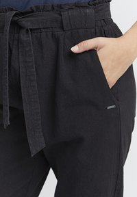 Black linen trousers featuring a high waistband with a tie, textured fabric, side pocket, and a small branded tag on the pocket.