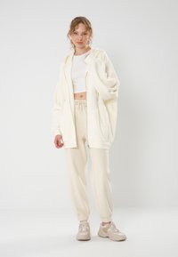 Hoodie crème surdimensionné avec fermeture éclair, associé à un pantalon de jogging crème avec poignets élastiques et taille à cordon, complété par des baskets beiges.