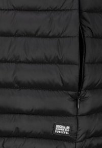 Schwarze Steppjacke mit horizontalen Nähten, teilweise offener Reißverschluss und einem schwarzen Patch mit der Aufschrift "ORIGINAL 86 ALWAYS BE BOLD SUBLEVEL."