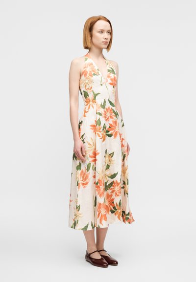 Φλοράλ φόρεμα με λαιμόκοψη halter σε κρέμ χρώμα, με πορτοκαλί και πράσινα ανθικά μοτίβα. Σιλουέτα Α με φούστα μεσαίου μήκους. Το μοντέλο φορά καφέ παπούτσια.