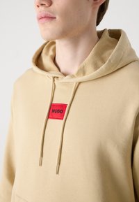 Sudadera beige con un parche rectangular rojo etiquetado "HUGO". Cuenta con capucha con cordón y forro interior texturizado. Tejido suave, corte relajado.