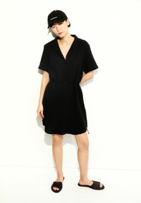 Schwarzes Hemdkleid mit kurzen Ärmeln, tailliertem Bund und lässiger Passform; kombiniert mit einer schwarzen Kappe und schwarzen Slipper-Sandalen.