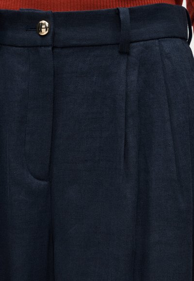 Παντελόνια σε σκούρο μπλε χρώμα με ψηλή μέση, θηλιές για ζώνη, πιέτες στο μπροστινό μέρος, πλαϊνές τσέπες και κλείσιμο με χρυσό κουμπί, συνδυασμένα με κόκκινο ριμπ πλεκτό πουλόβερ.