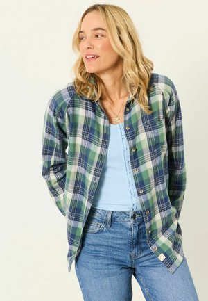 Femme aux cheveux blonds portant un débardeur bleu boutonné, une chemise à carreaux verts et bleus, et un jean bleu, regardant sur le côté.