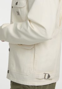 Helle cremefarbene_canvasjacke mit lockerer Passform. Sie verfügt über eine Brusttasche, metallene Knöpfe und einen verstellbaren Gurt am Saum.