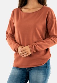 Donna che indossa una camicia a maniche lunghe color ruggine e jeans blu, con le mani conserte e un anello d'argento all'anulare sinistro su uno sfondo bianco.