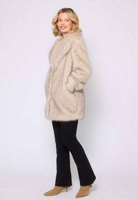 Faux fur-kappa i ljusbeige med en bred krage, sidofickor och en mjuk, fluffig textur. Matchas med svarta utsvängda byxor och beige stövletter.