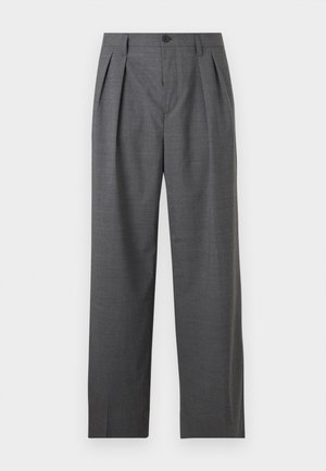 Homme portant un pull beige, un pantalon ample gris foncé, des sandales noires, tenant un sac noir devant un fond clair uni.