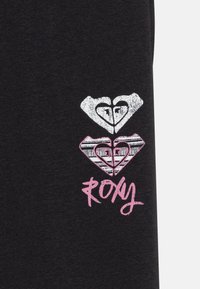 Svart tyg med ett grafiskt mönster av två överlappande hjärtan och ordet "ROXY" i rosa, med ett strukturerat mönster inuti hjärtana.