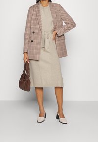 Blazer à carreaux avec double boutonnage, robe en maille beige avec ceinture nouée, sac à main marron, et chaussures à talons blocs beiges avec semelles noires.