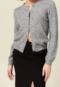 Jasnoszary sweter na drutach z okrągłym dekoltem, długimi rękawami i zapięciem na guziki. Posiada teksturowany wzór oraz ściągacze na mankietach.