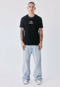 Zwarte katoenen t-shirt met PXP-logo, gecombineerd met lichtblauwe wijde jeans. Model draagt witte sneakers en een zilveren kettinghalsketting.